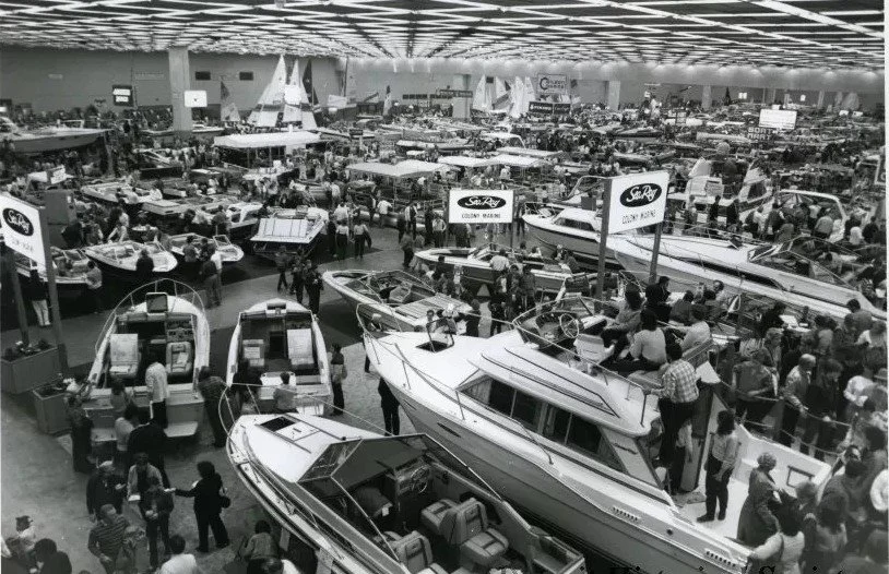 Indoor boat show with various boats displayed and many visitors walking around and examining them. Sea Ray Boats display is prominent.