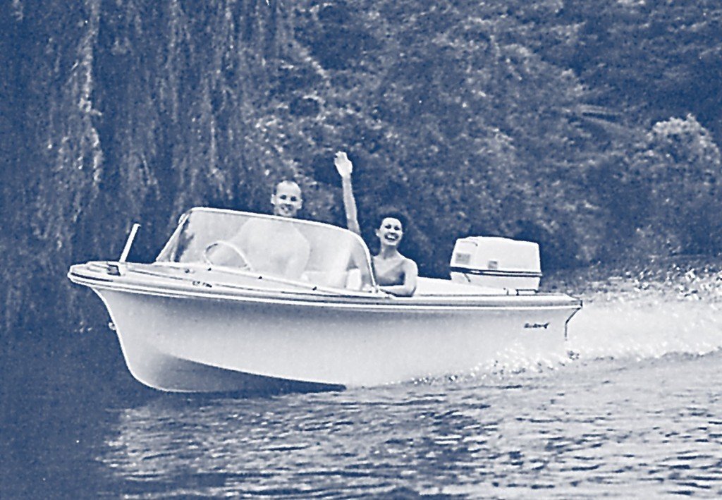 Two people, a man and a woman, are riding in a small motorboat on a body of water, with the woman waving. The boat has a canopy and an outboard motor, and there are trees in the background. An original Sea Ray boat.