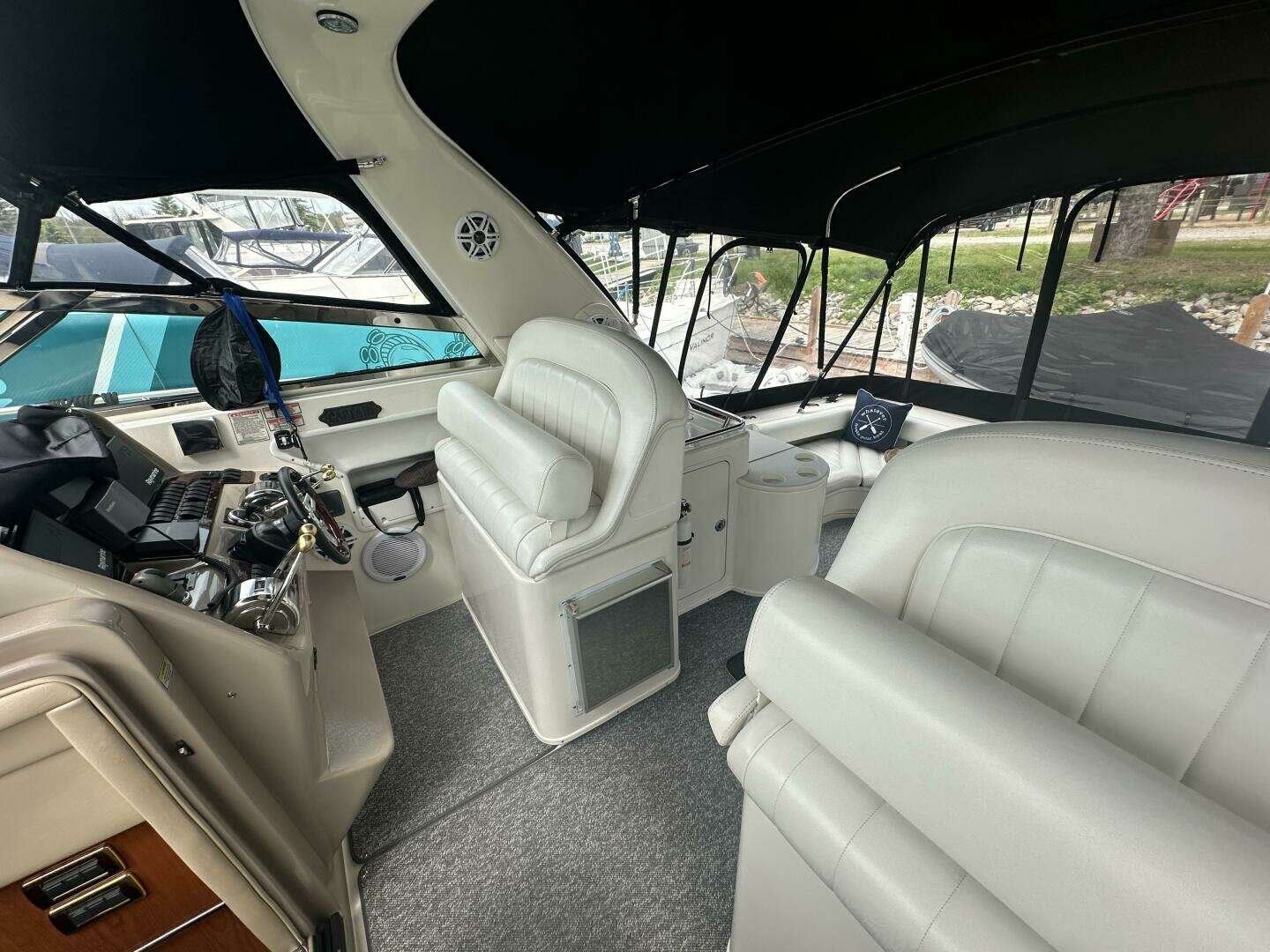 Interior view of a boat with white leather seating, a steering console, and large windows showing boats and water outside.