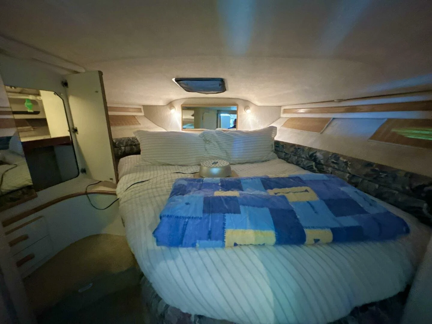 A cozy Sea Ray boat's bedroom with a round bed, striped pillows, a patchwork quilt, a small window, and built-in shelves along the walls.