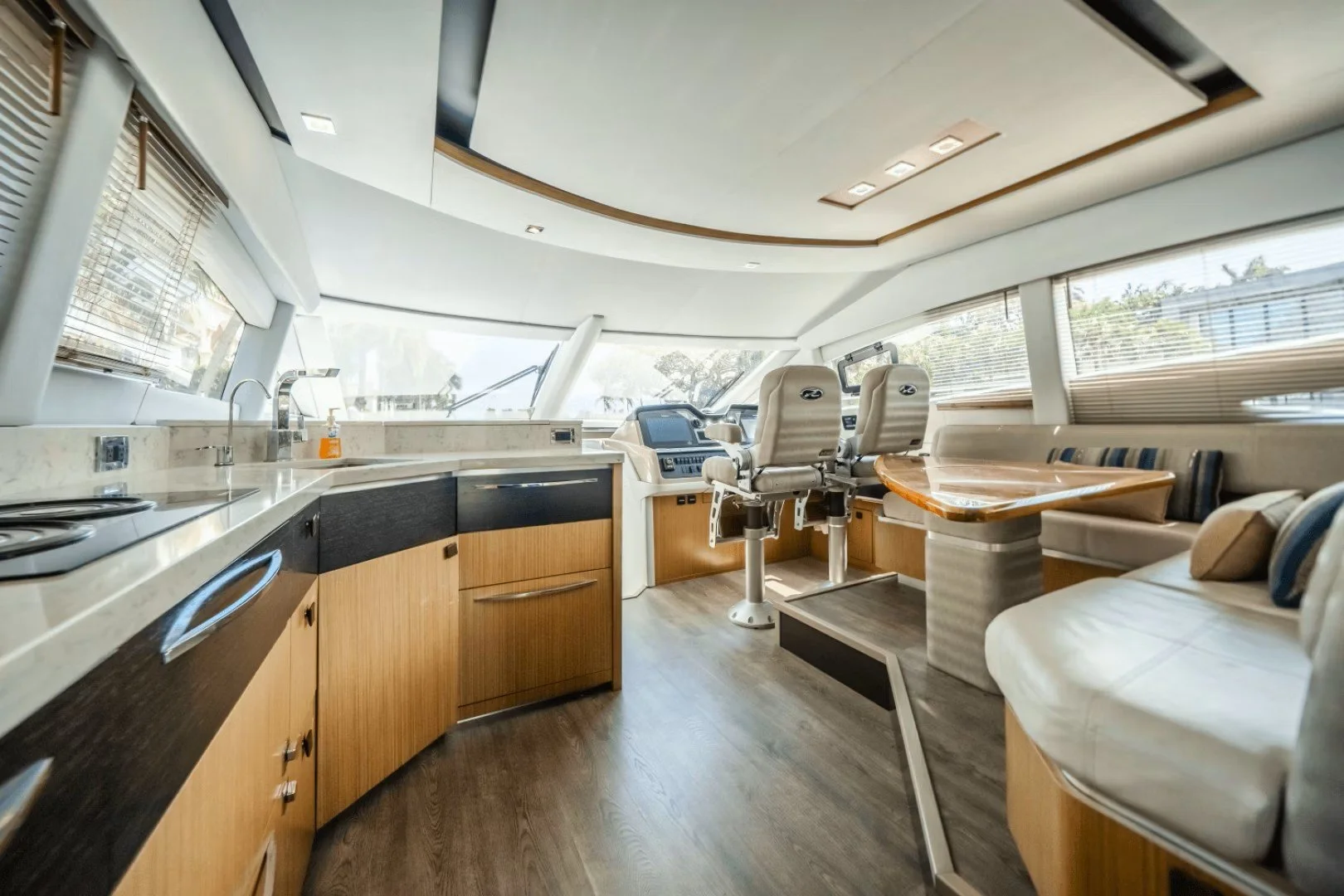 Interior of a yacht with a kitchen area, captain's chair, and seating area with cushions, large windows with blinds, and wood flooring.