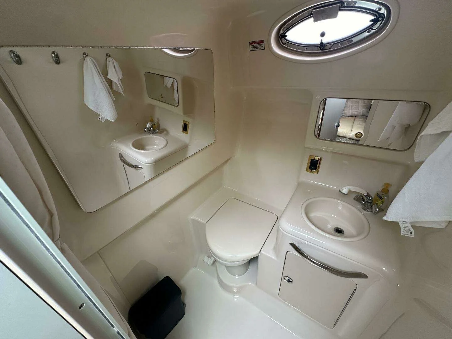 Boa bathroom with a toilet, a compact sink with a curved handle, a mirror, a small oval window with a latch, and a white towel hanging on the side. The walls and fixtures are cream-colored with a smooth, shiny finish.
