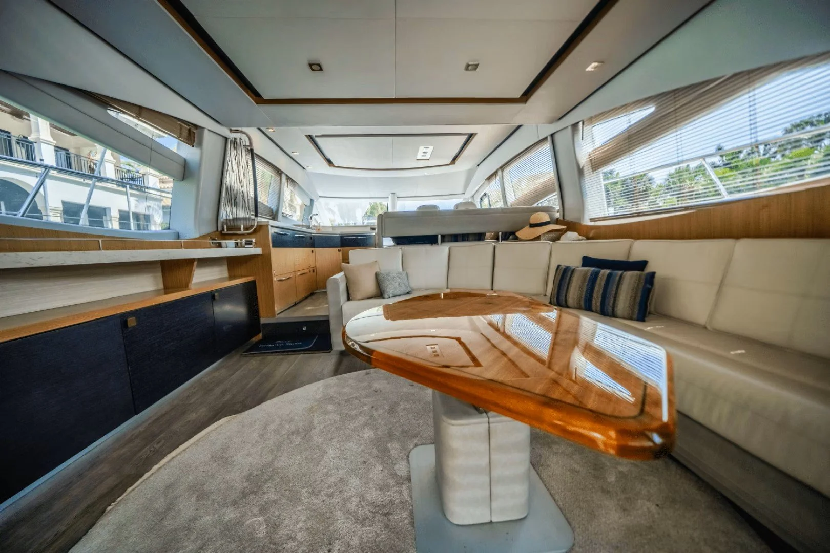 Luxury interior of a yacht with a cream sectional sofa, a wooden coffee table, large windows with blinds, and a storage area with beige cupboards.