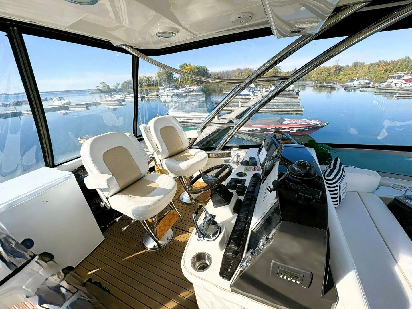 View of a boat's enclosed cockpit with white captain and passenger chairs, steering wheel, and control panel, overlooking a marina with parked boats, calm water, and trees in the distance.
