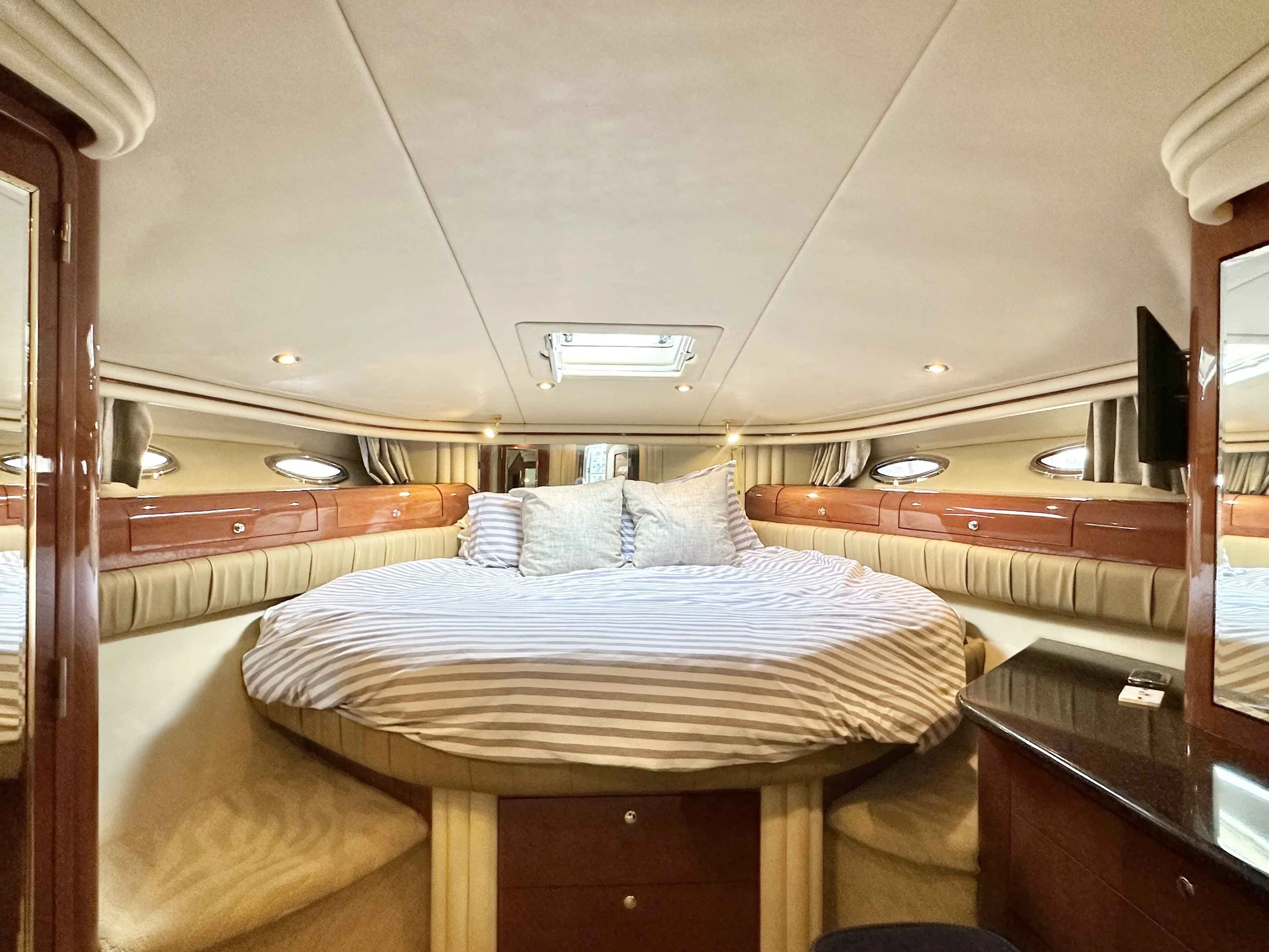 Luxurious Sea Ray yacht's bedroom with a rounded bed, striped bedding, and multiple pillows, small windows, overhead skylight, and wood accents.