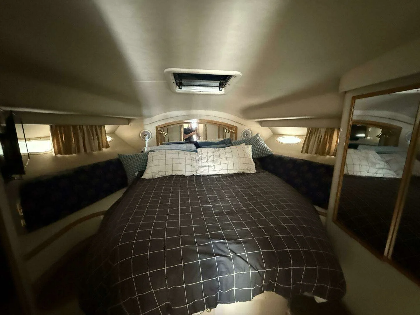 Interior view of a cozy boat cabin with a bed, multiple pillows, small windows, and a mirror on the wall.