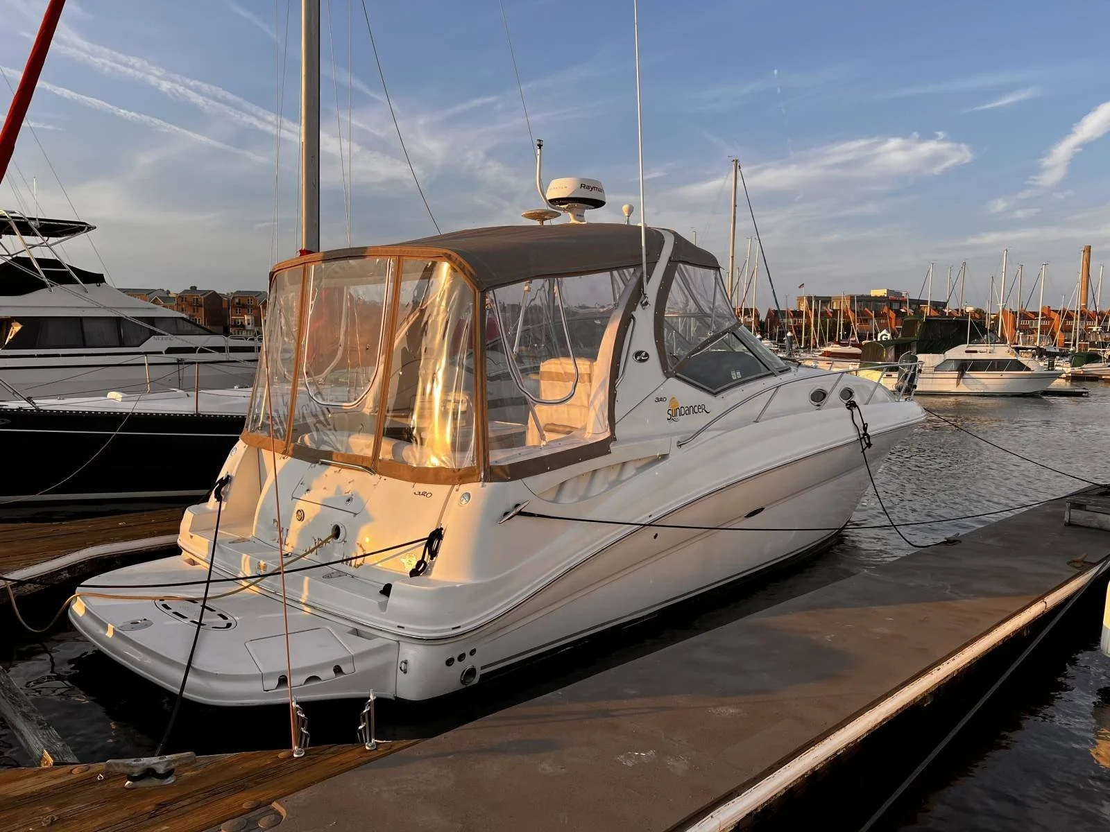 A white Sea Ray 320 Sundancer motorboat docked at a marina, with other boats and buildings in the background, under a calm early evening summer cloudy sky.