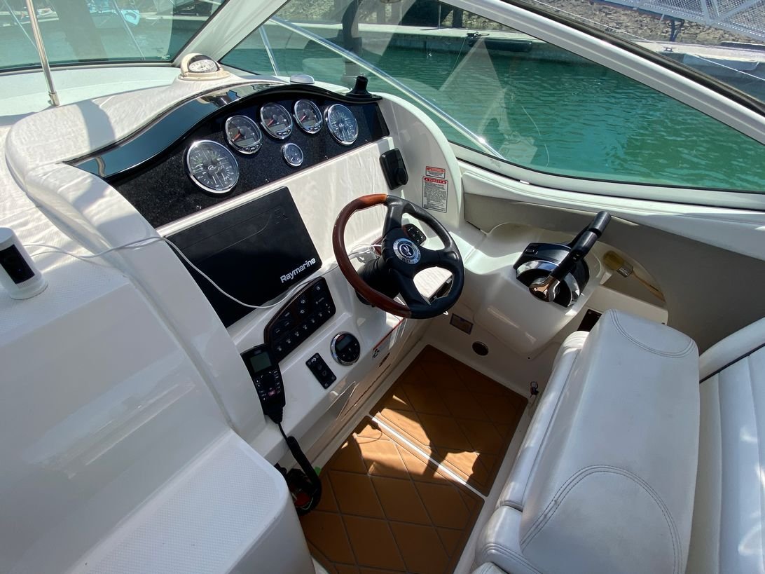 Interior of a yacht's helm station showing a steering wheel, an array of gauges, a Raymarine navigation display, throttles, and seating, with water visible through the front windows.