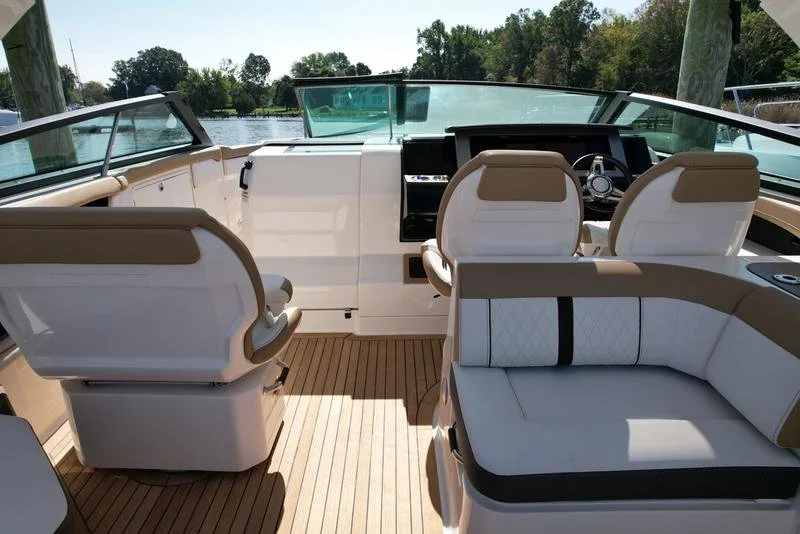 View of the helm and seating area on a boat with beige and white upholstery, situated near a body of water with trees and blue sky in the background.