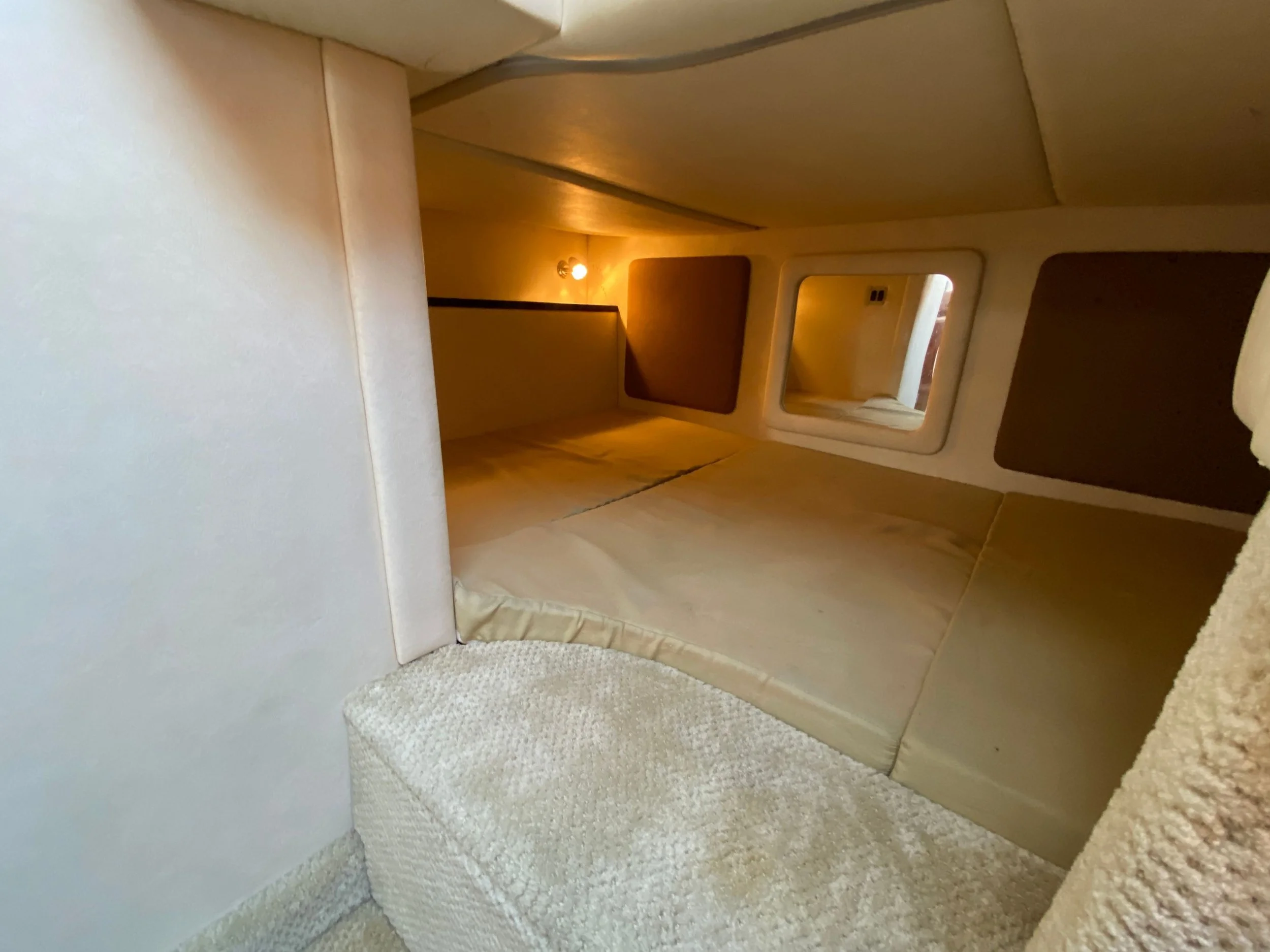 Interior of a boat's sleeping area with a bed, small window, plush beige cushions, and soft lighting.