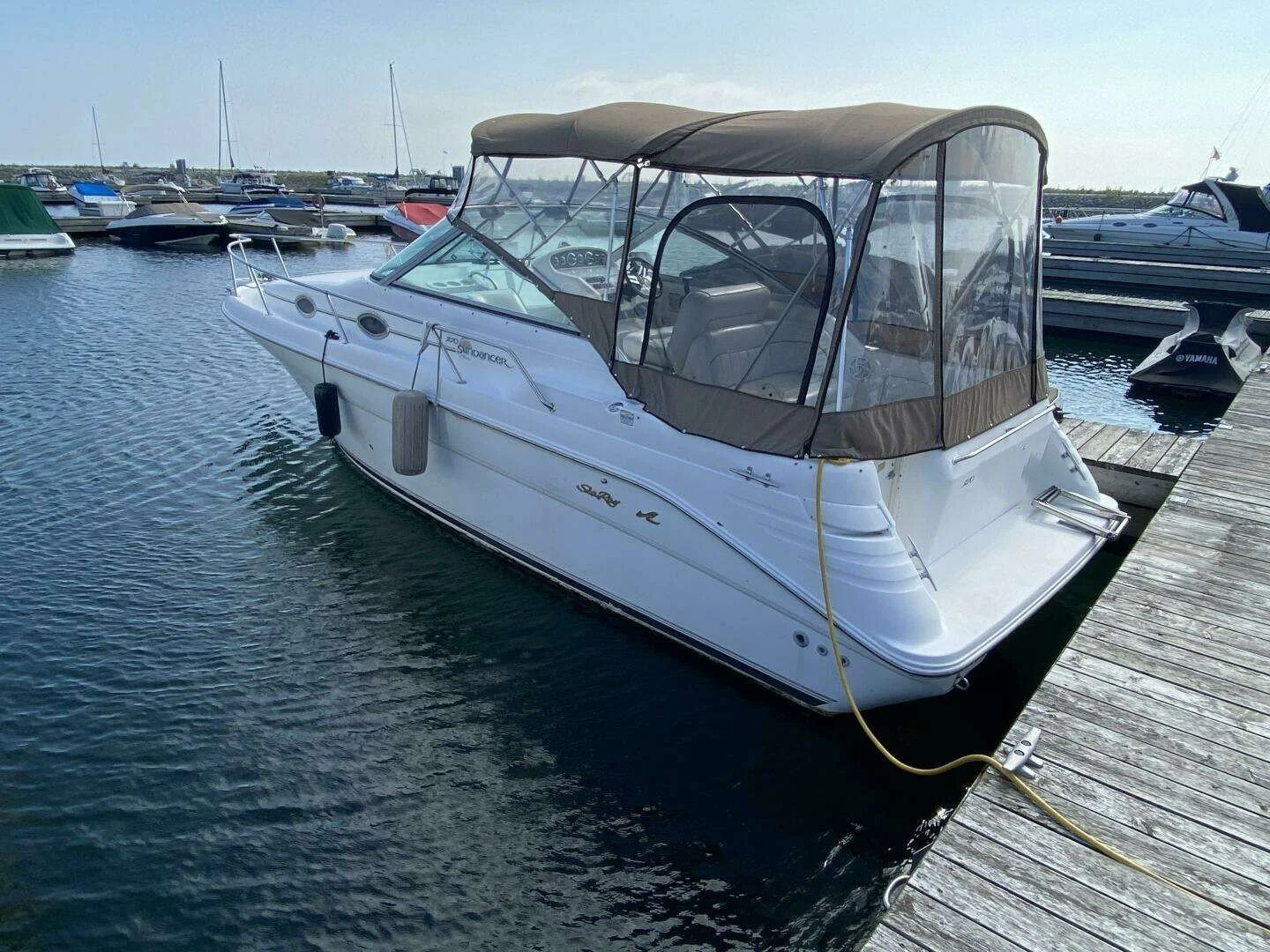 A white express style boat docked at a marina with a brown canvas canopy and clear plastic windows. Several other boats are visible in the water and on the docks in the background. Boat is a Sea Ray 270 Sundancer.