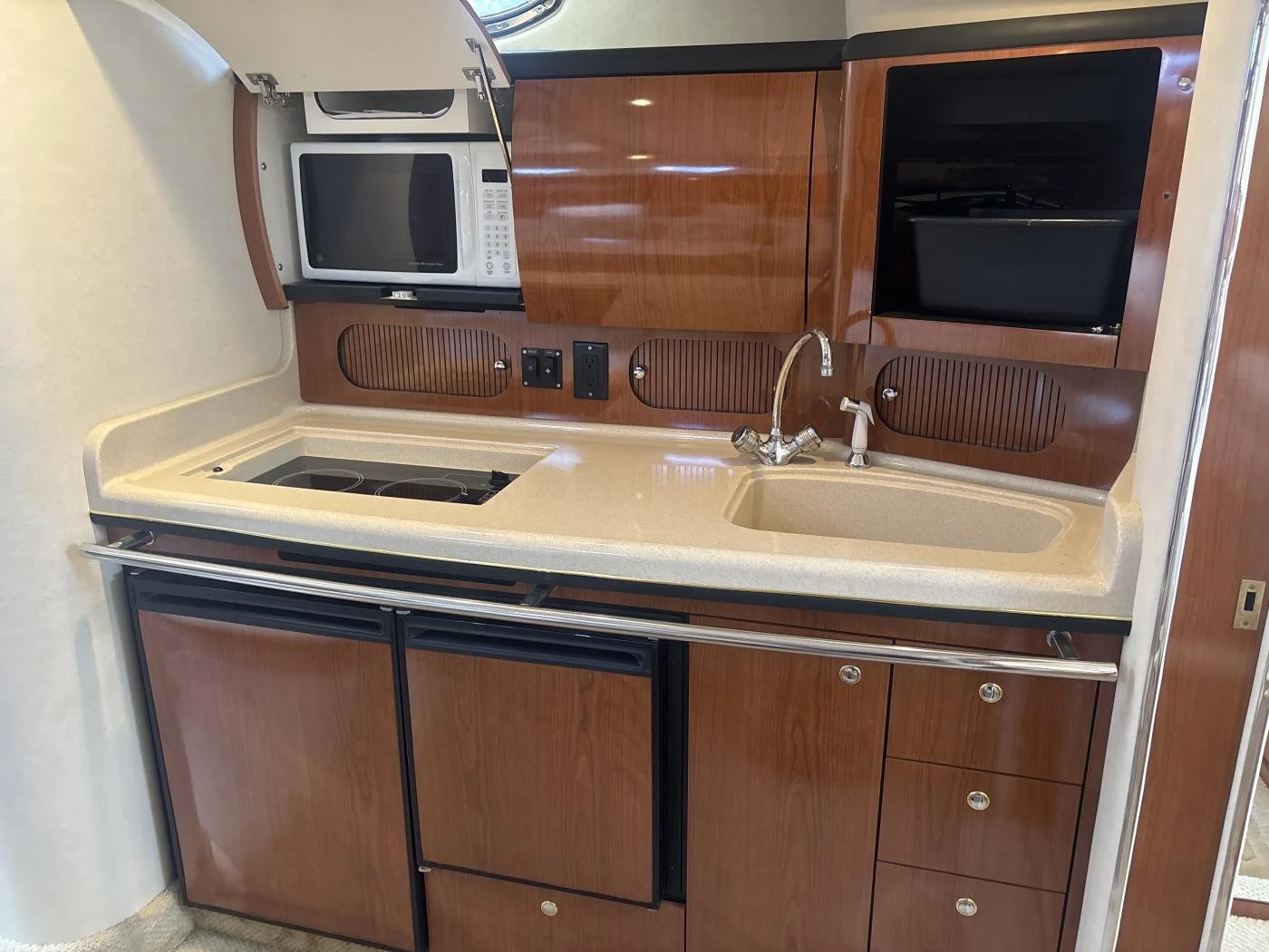 A small kitchen galley area on a Sea Ray Sundancer boat, with cherry wood cabinets, a beige countertop, a built-in sink with a faucet, a microwave, a stovetop, and an open cabinet with a toaster inside.