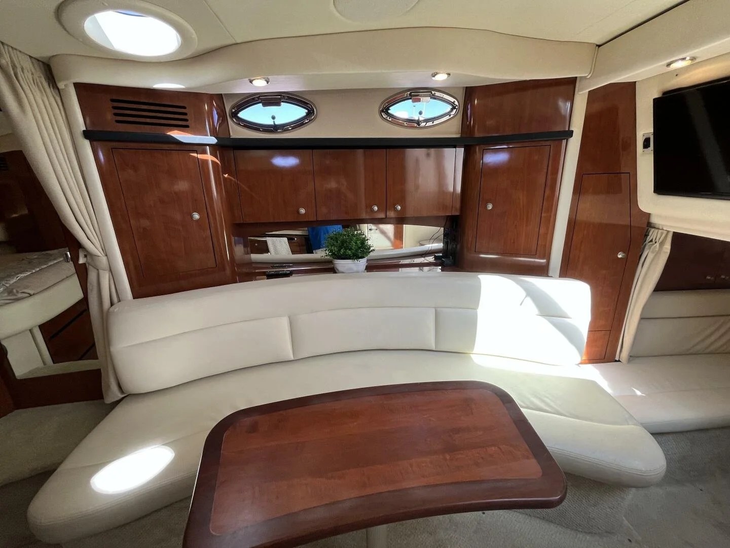 Interior of a luxury Sea Ray 340 Sundancer yacht with cream-colored cushioned seating, a wooden table, wood-paneled cabinets, oval portholes, and a wall-mounted TV.
