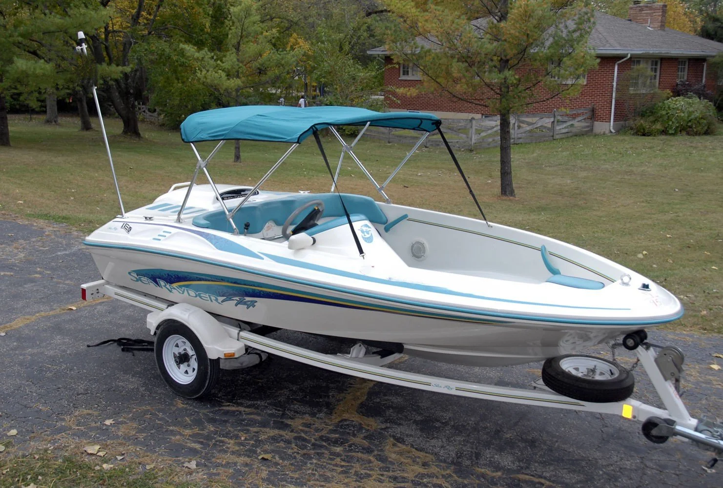 White and blue speedboat on a trailer parked on a paved lot with grass, trees, and a brick house in the background. Sea Ray F-14 Sea Rayder.