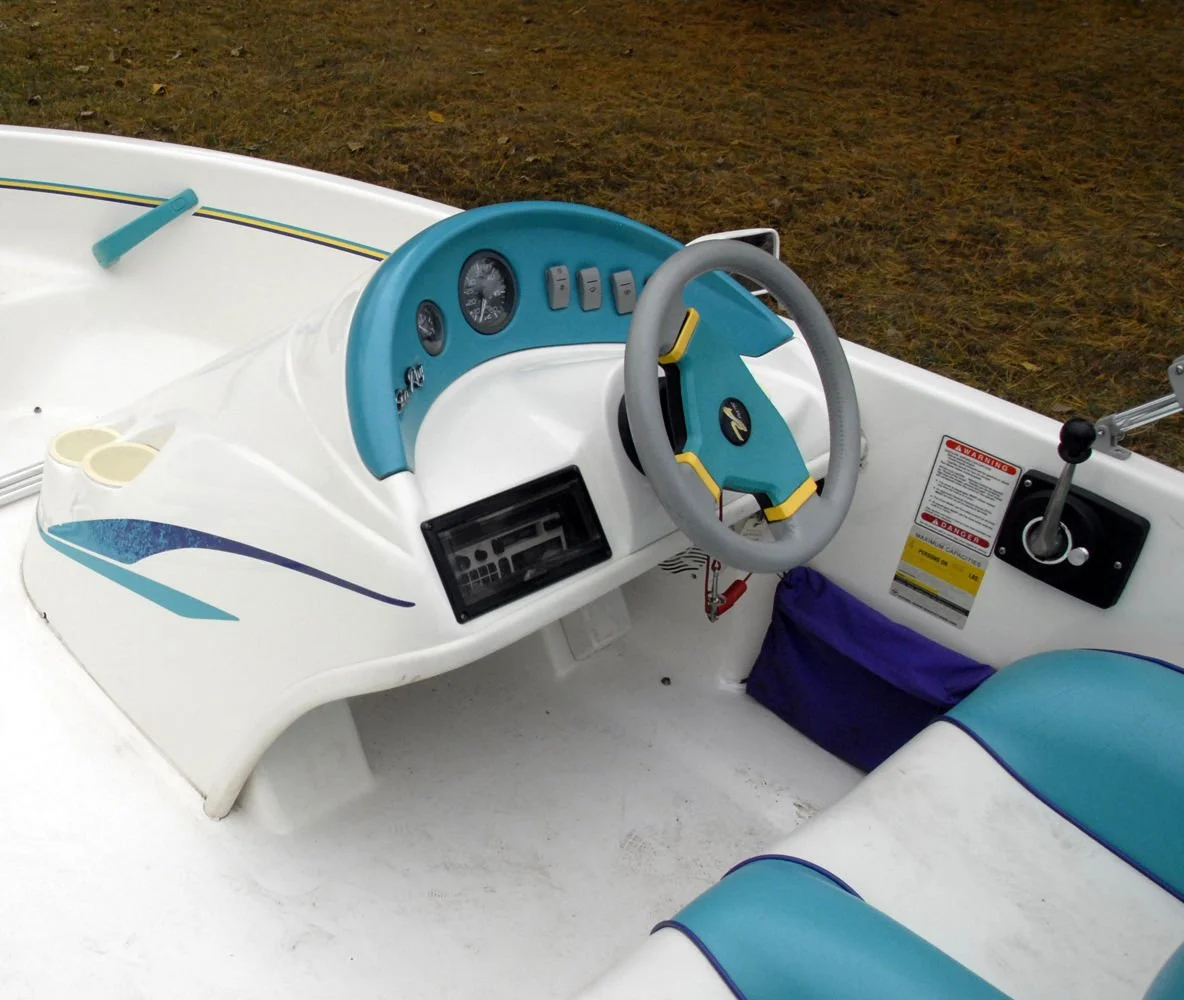 The interior of a small boat with a steering wheel, dashboard, and control switches, on a grassy area.
