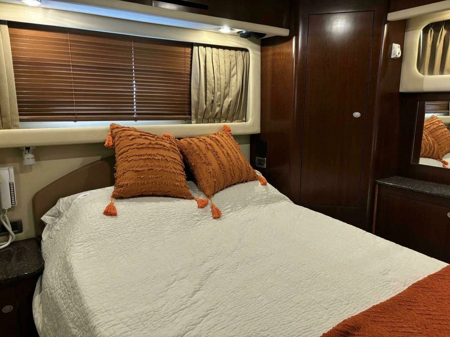 A Sea Ray yacht's bedroom with a bed that has white bedding and two orange pillows with tassels. There are wooden blinds on a window and dark wood cabinets, with a small mirror and a black countertop.