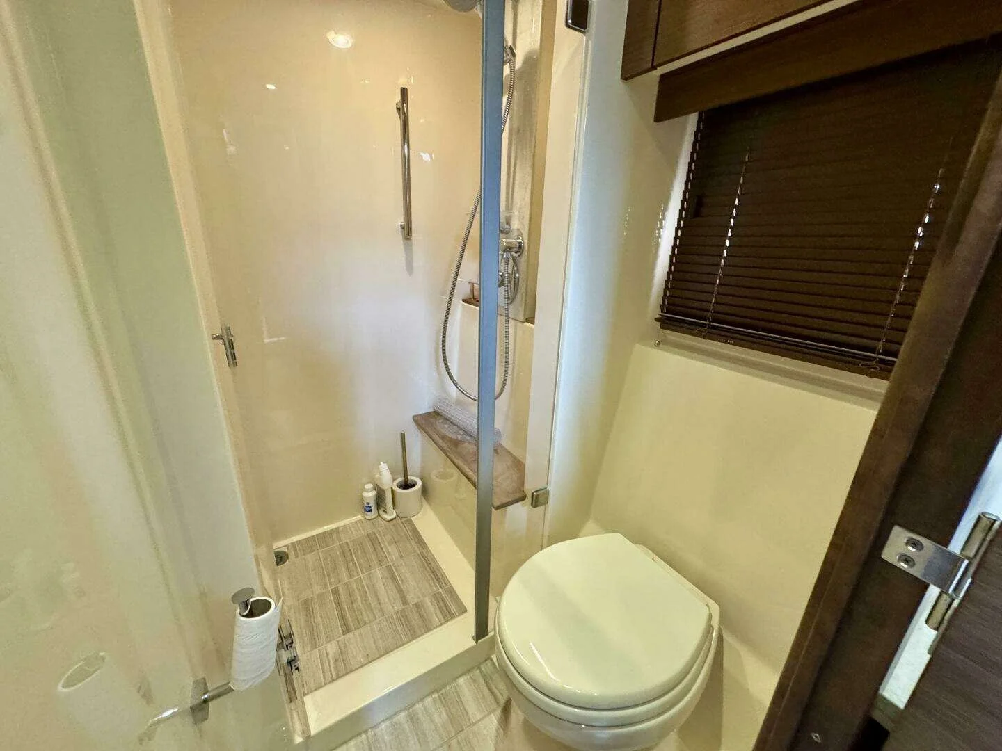 Marine bathroom / head with a toilet next to a small enclosed shower area with a glass door, shower head, and toiletries, and a window with closed dark blinds. Sea Ray yacht.
