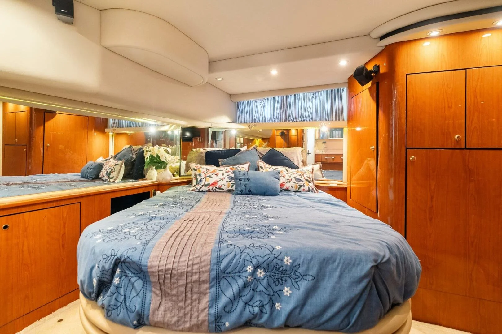 A boat's bedroom with a large bed covered in blue and gray bedding, decorative pillows, and a mirror running along one wall. There are wood cabinets and a window with striped curtains, and the room has warm lighting. Sea Ray yacht.