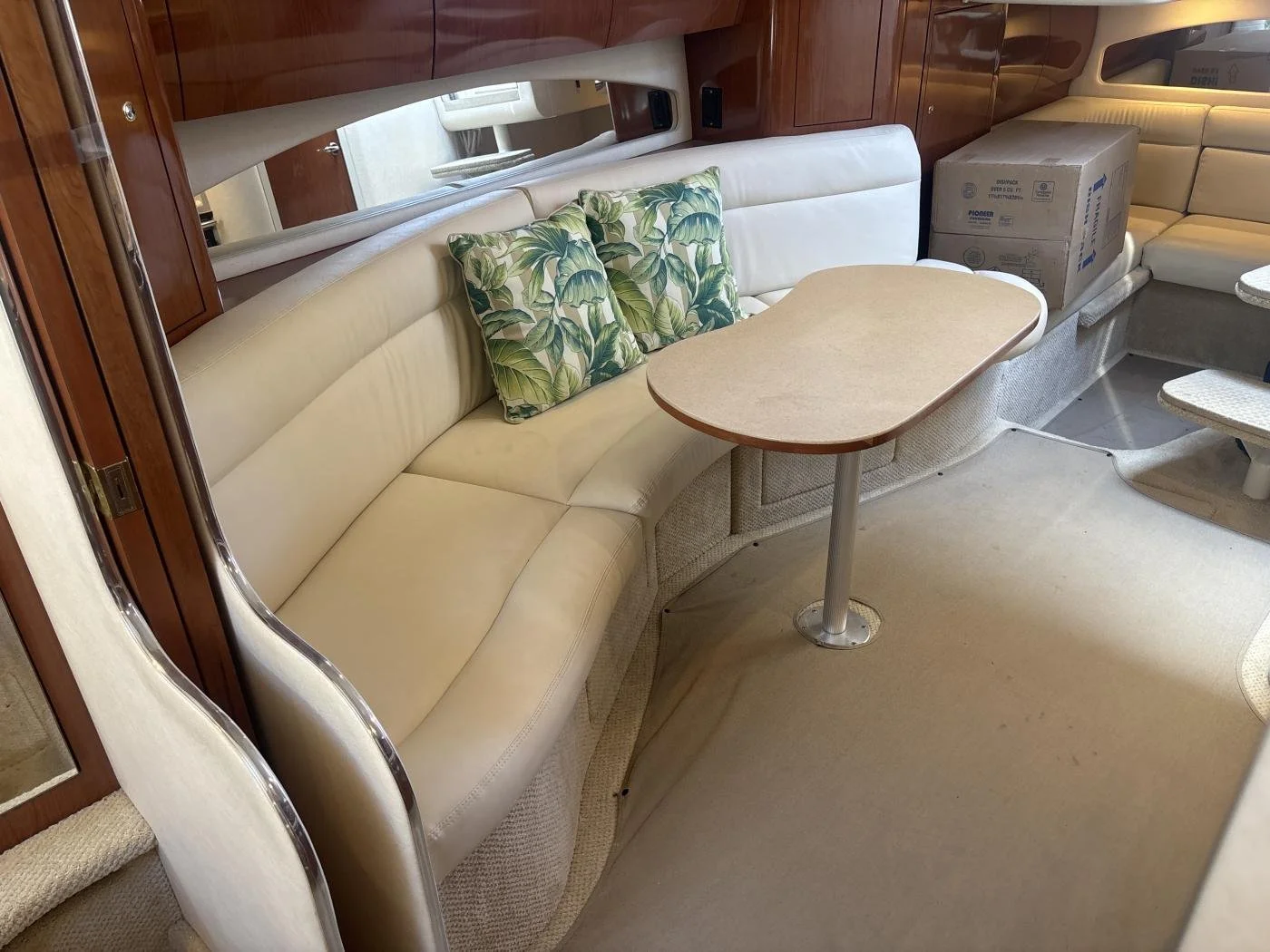 Interior of a Sea Ray boat with a beige curved sofa, two tropical print pillows, a small oval table, and a cardboard box in the background.
