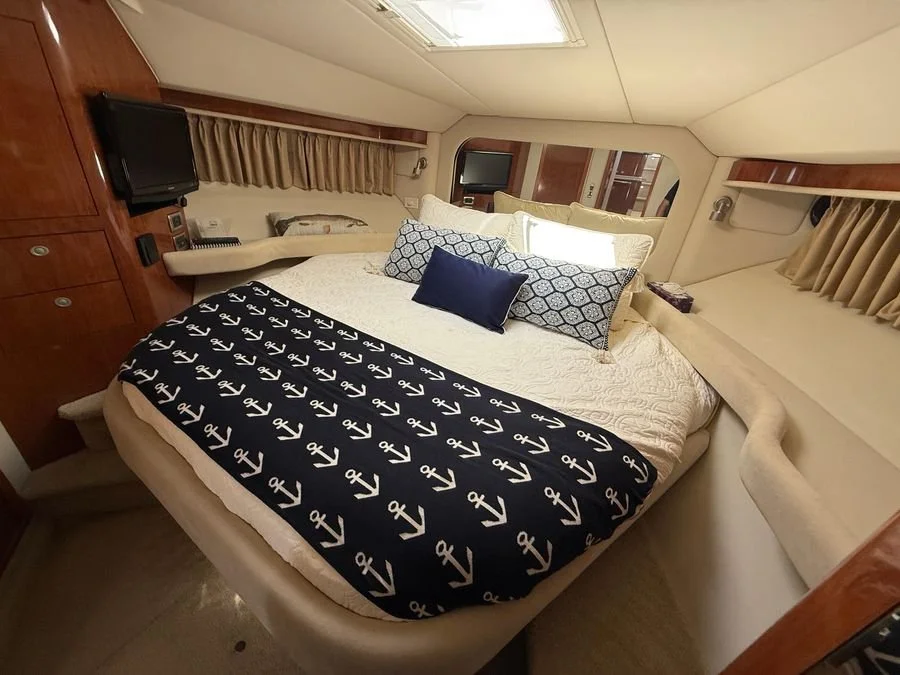 A boat's luxury bedroom with a bed covered in nautical-themed pillows and blanket, small wall-mounted televisions, and beige curtains in a Sea Ray yacht interior.