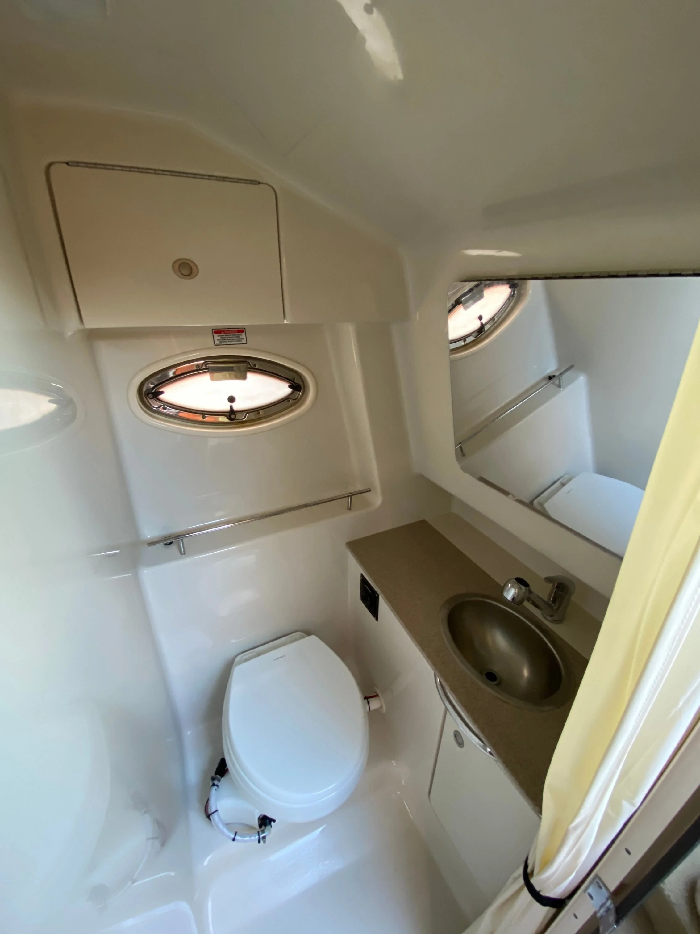 A boat's bathroom with a toilet, a small sink with a countertop, a mirror above the sink, an oval port-light window.