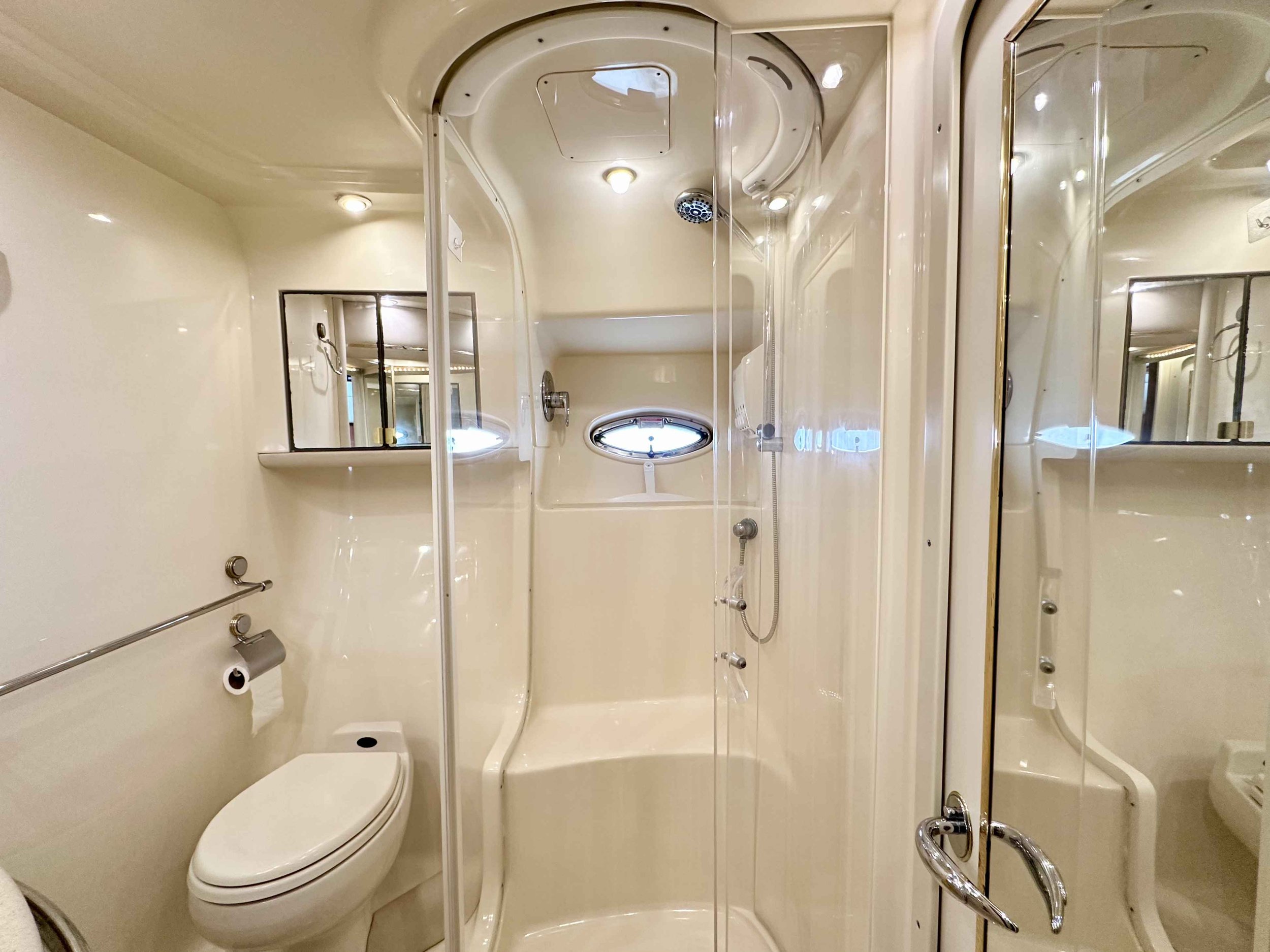 A Sea Ray yacht's enclosed bathroom with a modern design, featuring a toilet, a curved shower with a hand-held showerhead, a small window, mirrors, and minimal fixtures.