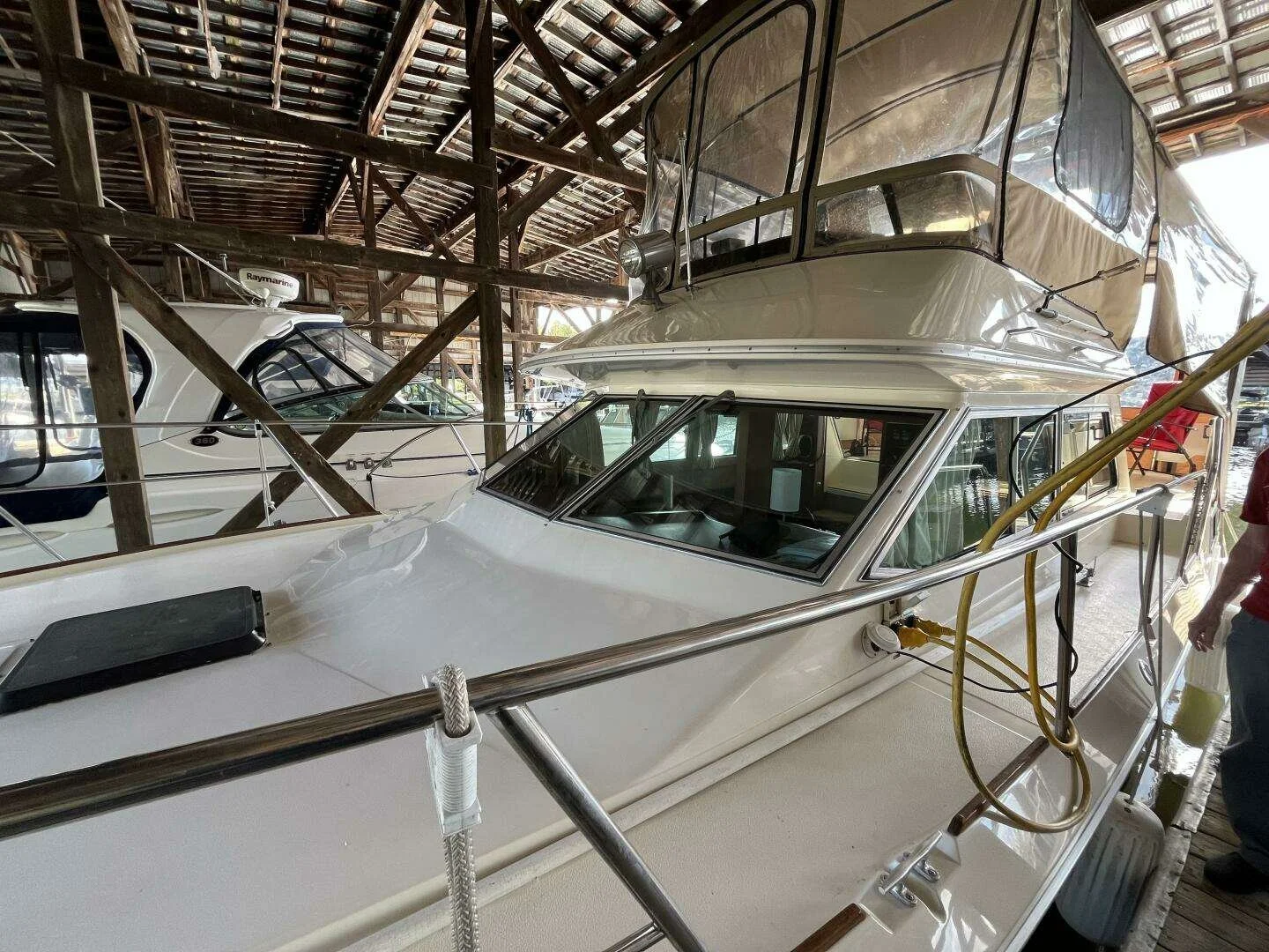 White yacht docked in a marina storage facility with a wooden roof, another boat visible in the background, and a person partially visible on the right side.