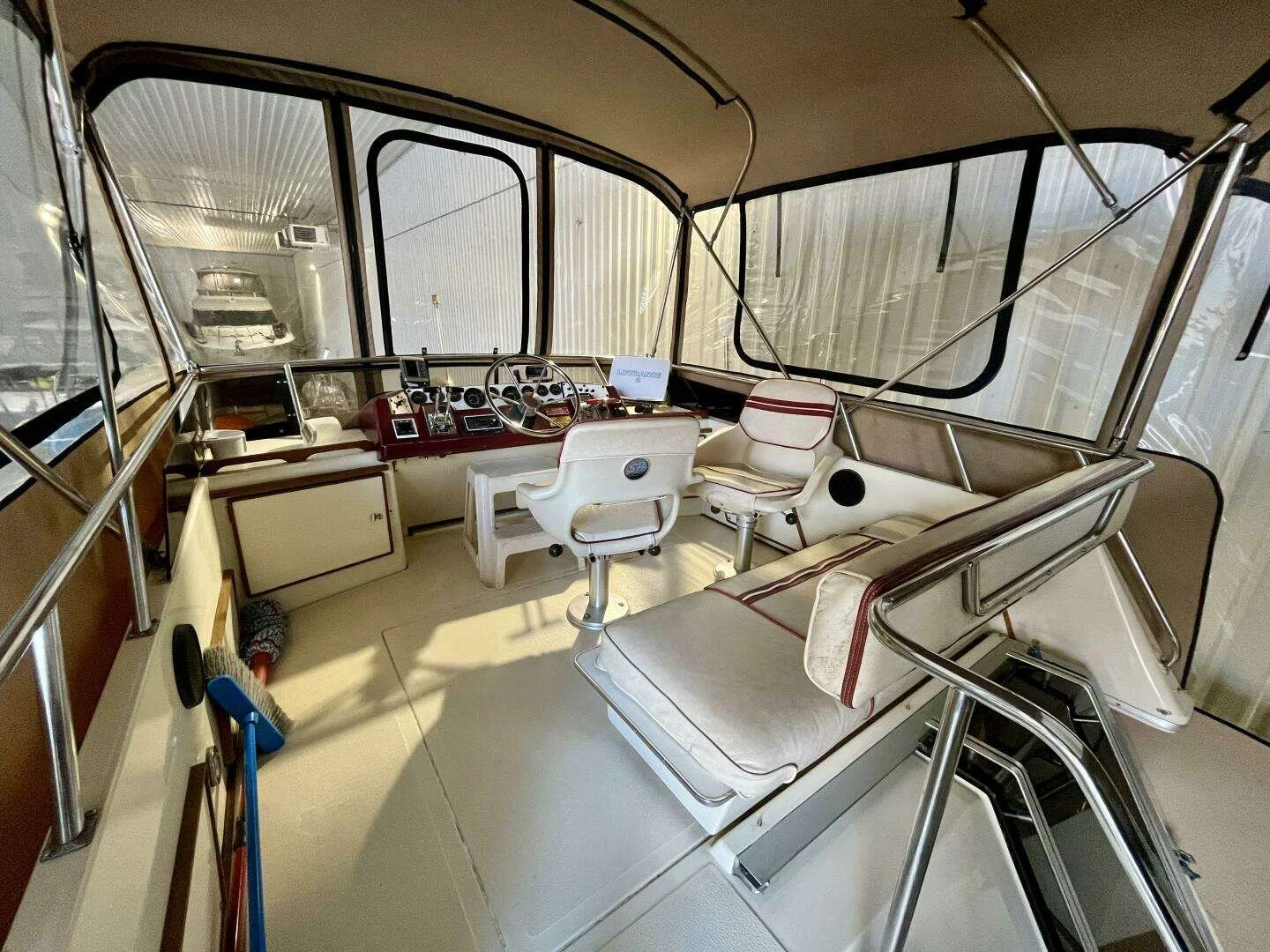Interior of a boat cabin with white and red cushioned seating, a steering wheel, and navigation equipment, surrounded by large windows.