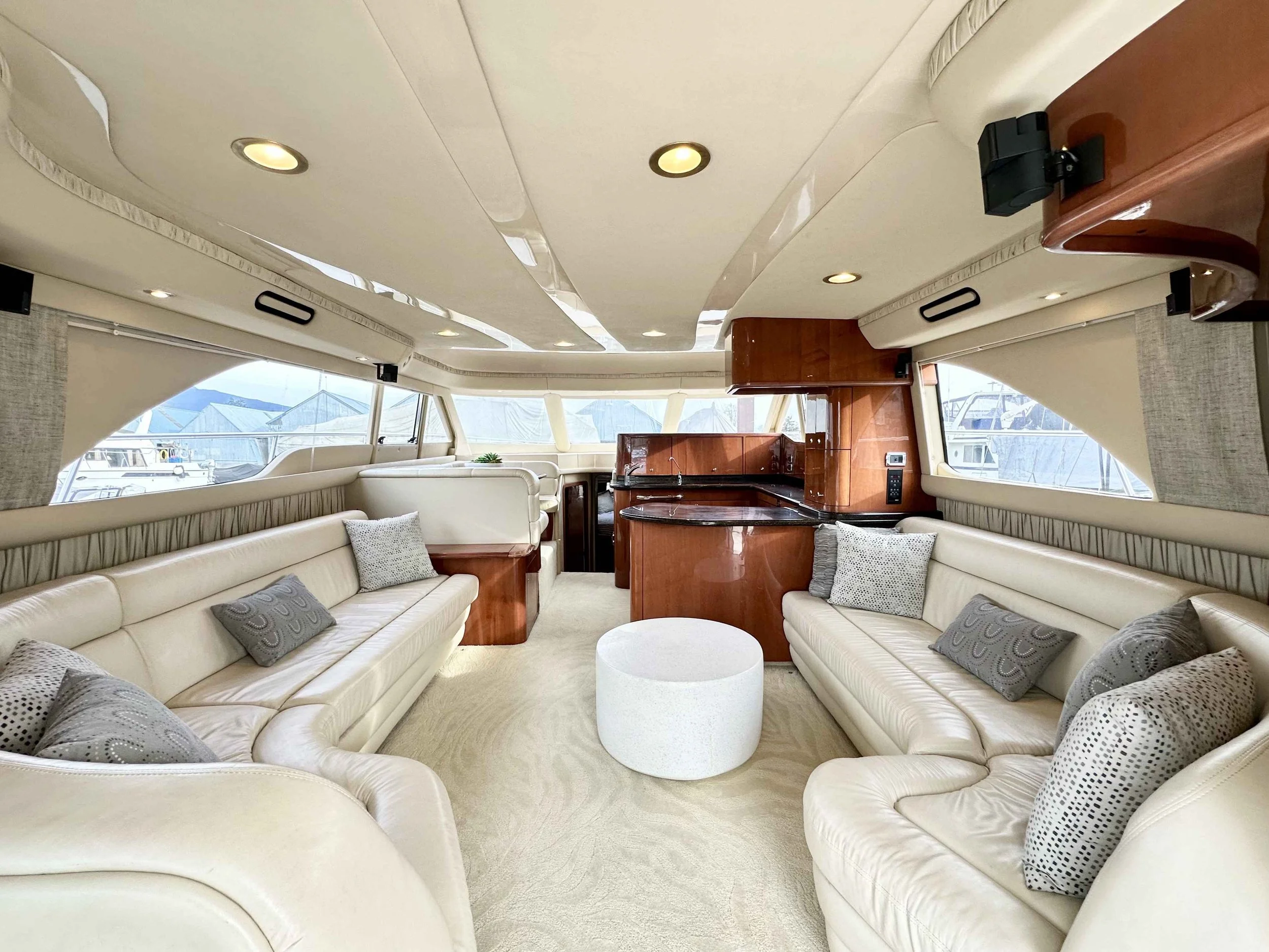Interior of a yacht's lounge area with cream-colored leather sofas, gray decorative pillows, and a small round white ottoman, with large windows showing boats outside.