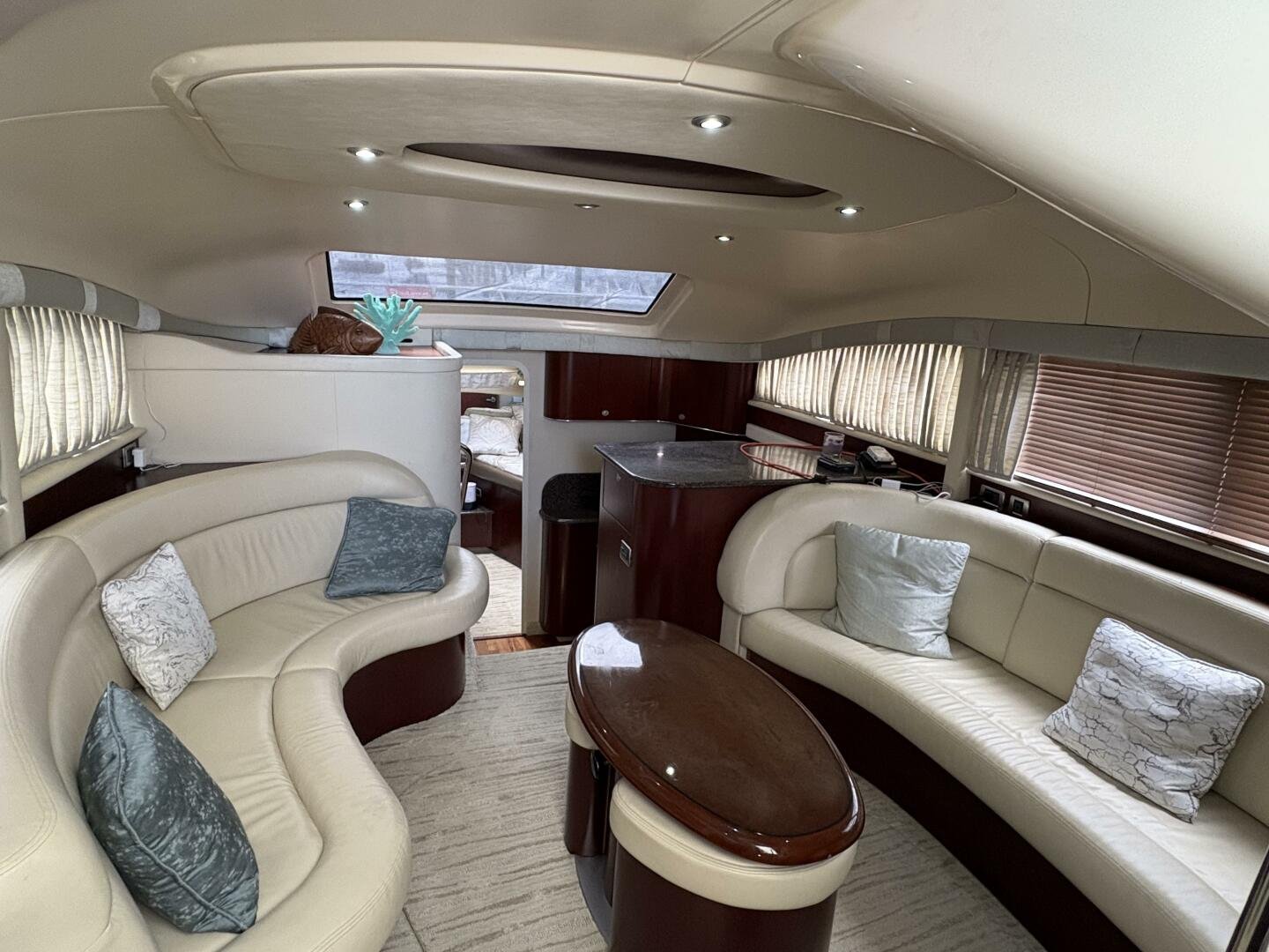 Interior of a yacht living area with curved cream leather couches, decorative pillows, a wooden coffee table, small kitchen area with granite countertop, windows with blinds, and a bed visible in the background.