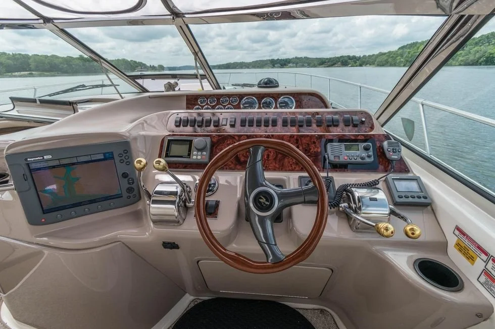 Interior view of a boat's helm station with navigation and control equipment, overlooking a body of water and lush green shoreline.