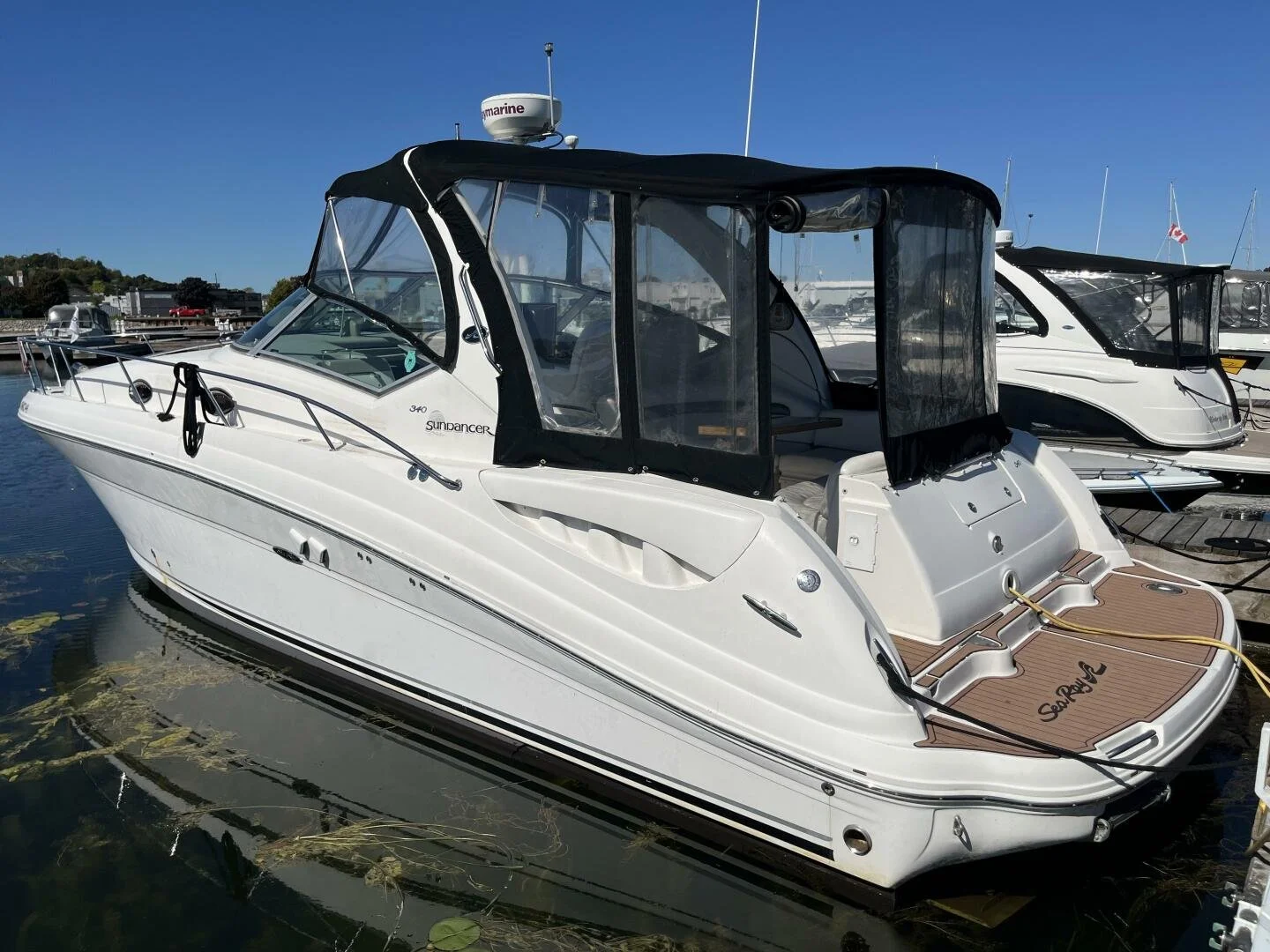 White motor yacht is docked at a marina, with black canvas covers. There are other boats in the background under a clear blue sky. Sea Ray 340 Sundancer, with sea deck custom marine flooring on swim platform.