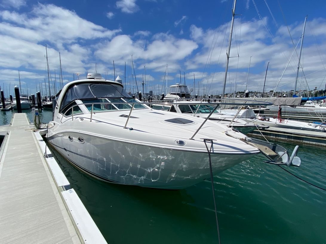 A luxury white yacht docked at a marina with other boats and sailboats in the background under a partly cloudy sky. Focus is on a Sea Ray 325 Sundancer.