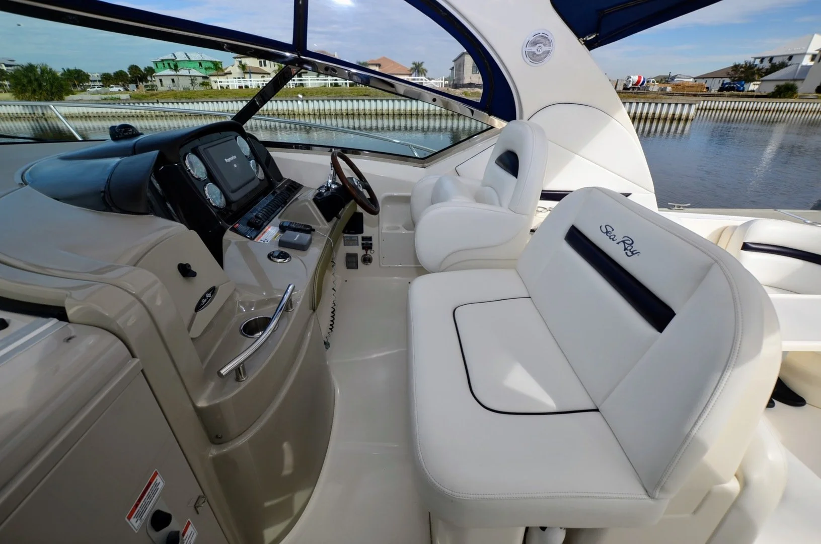 Interior of a Sea Ray yacht's cockpit showing white leather seats, steering wheel, dashboard with controls, and view of water outside.