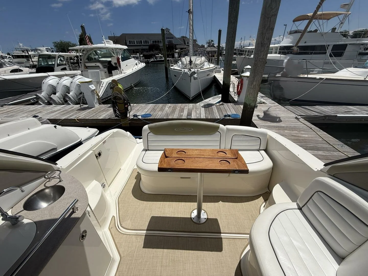 View from the deck of a boat docked at a marina, showing seating area with white cushioned benches, a small wooden table, and a sink. Several boats are docked adjacent, with a wooden pier and maritime equipment visible.