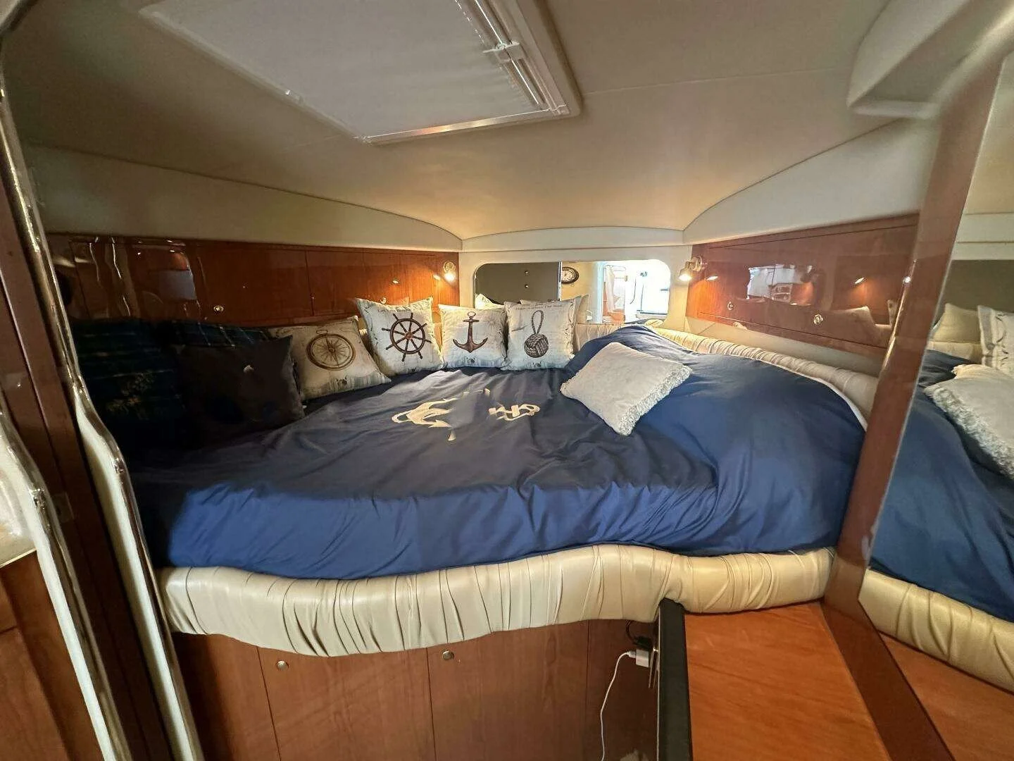 Interior of a boat cabin with a large bed, navy blue bedding, decorative pillows with nautical designs like an anchor, steering wheel, and anchor chain, wood cabinetry, small lights.