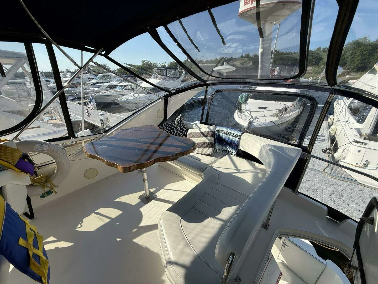 Interior view of a boat with a small wooden table, white cushioned seating, and several pillows. Large windows show other boats docked at a marina in the background.