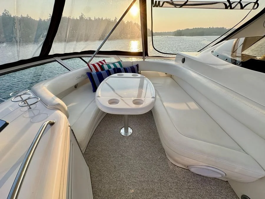 Interior of a boat with a white cushioned seating area, small round table with built-in cup holders, colorful pillows, and a view of water and sunset through the windows. Flybridge seating on Sea Ray 480 Sedan Bridge boat.