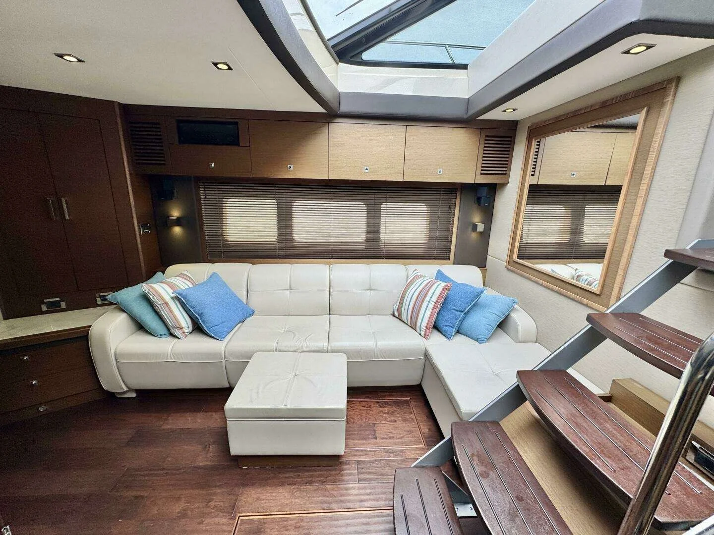 Interior of a yacht's living room with a white sectional sofa with blue and striped pillows, a small white ottoman, wooden stairs on the right, a large mirror on the right wall, wooden cabinets, and skylights in the ceiling. Sea Ray boat.