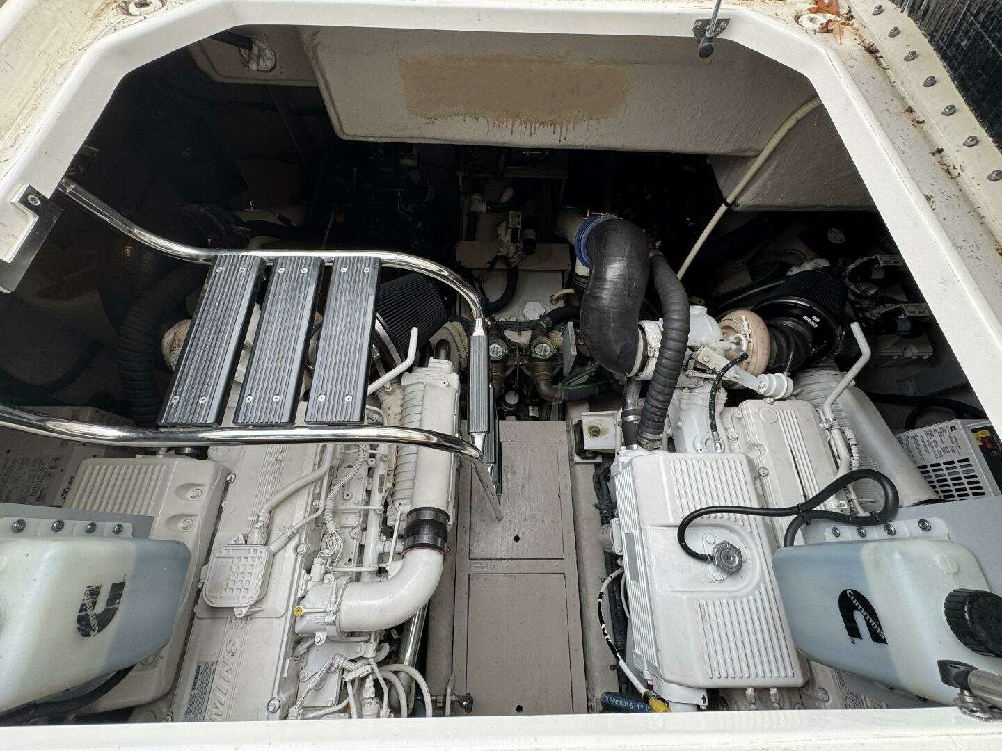 Interior view of a boat engine compartment showing two white Cummins diesel engines, black hoses, and metal components.