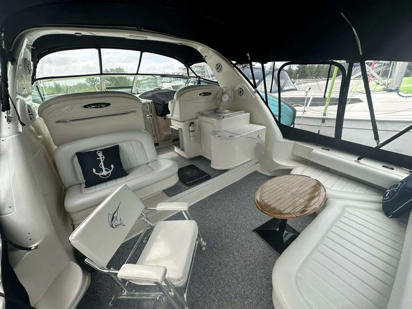 Interior of a yacht with white cushioned seating, a small wooden table, two white chairs, a navy pillow with an anchor, and a fan, overlooking a marina with other boats.