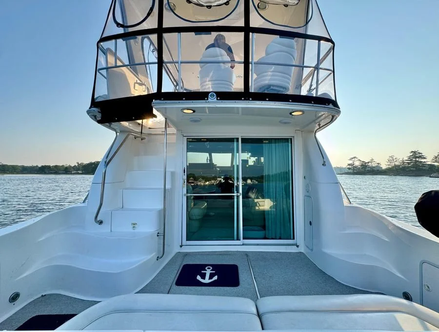 View of the deck of a white yacht with sliding glass doors leading inside, and upper deck with chairs and a person inside, on the water with a shoreline and trees in the background. Cockpit of a Sea Ray 480 Sedan Bridge boat.