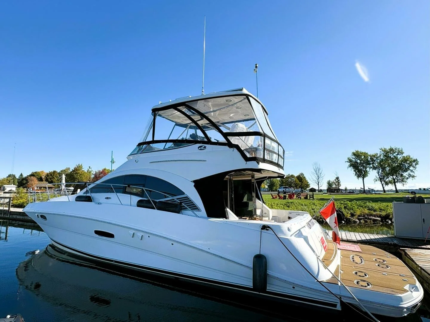 A white Sea Ray yacht docked at a marina on a bright, clear and sunny summer day with blue skies and trees in the background. Boat is a 47 Sedan Bridge model.