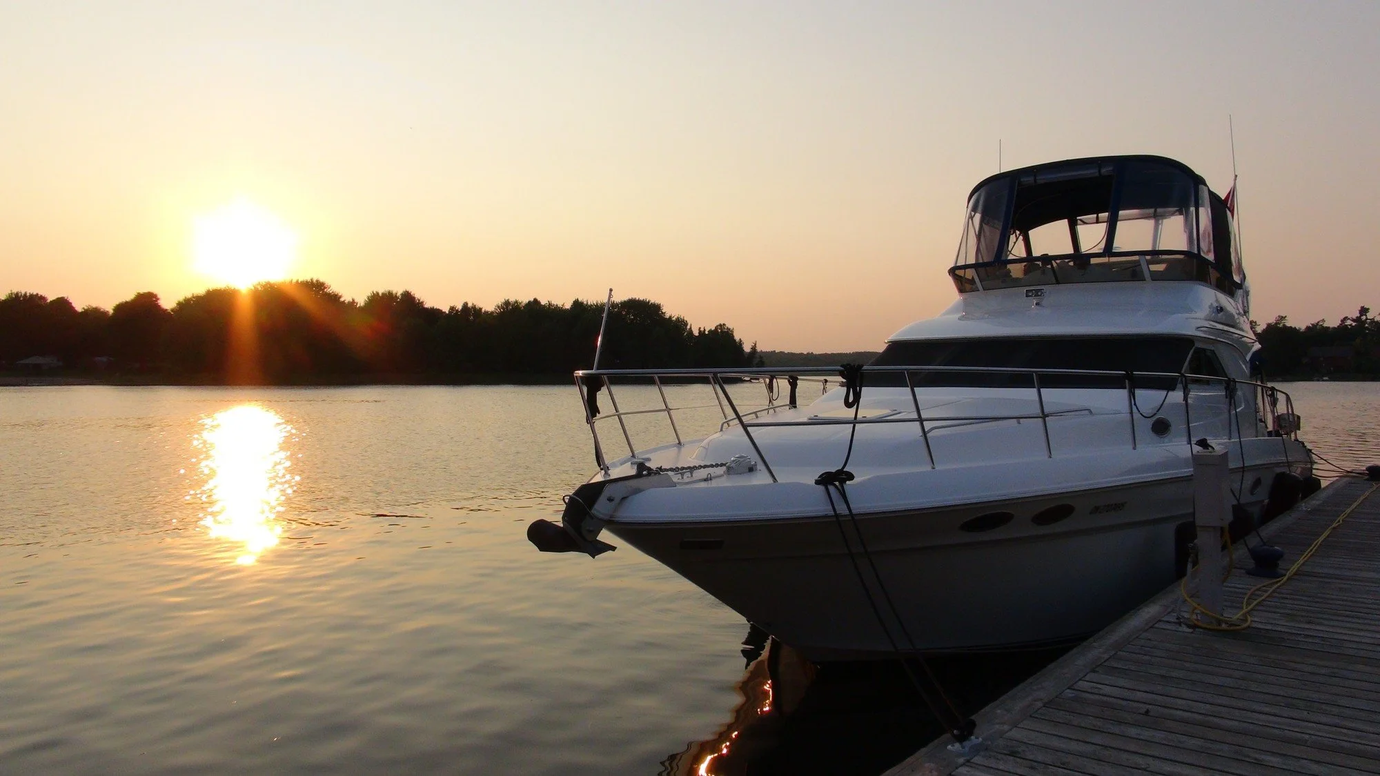 A yacht docked at a pier during sunset, with calm water reflecting the sunlight and trees in the background. Sea Ray 400 Sedan Bridge boat.