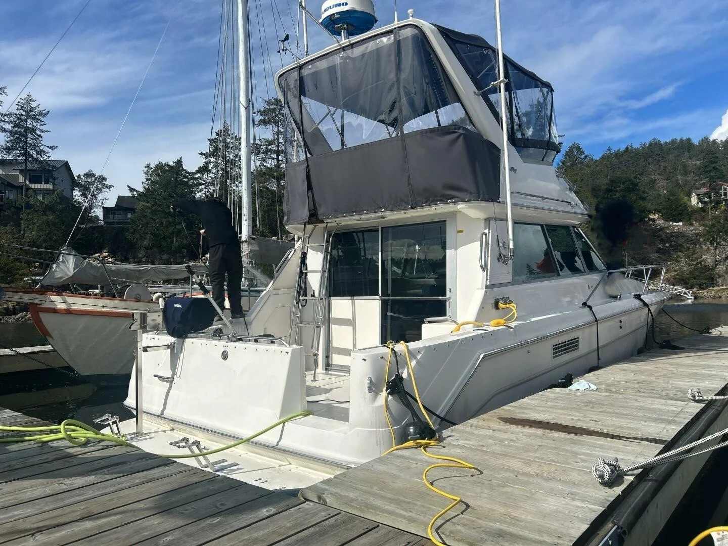 A white yacht docked at a wooden pier. The yacht has a covered upper deck and is connected to power and water supplies by yellow and green hoses. The background shows a hillside with houses and trees under a partly cloudy blue sky.