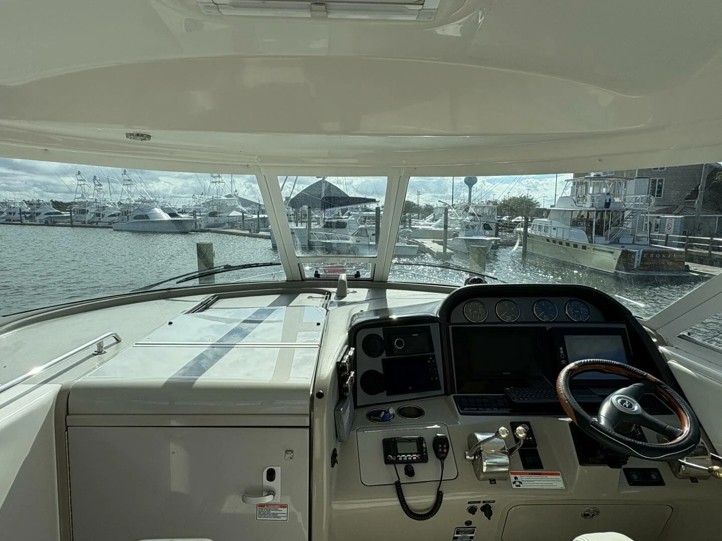View from inside a boat's cabin showing the steering wheel, control panels, and navigation instruments, with a marina filled with docked boats visible through the front windshield.