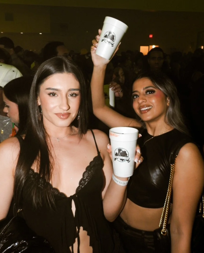 Two women at a crowded indoor party holding large white cups with black logos, one woman is winking and the other is smiling.