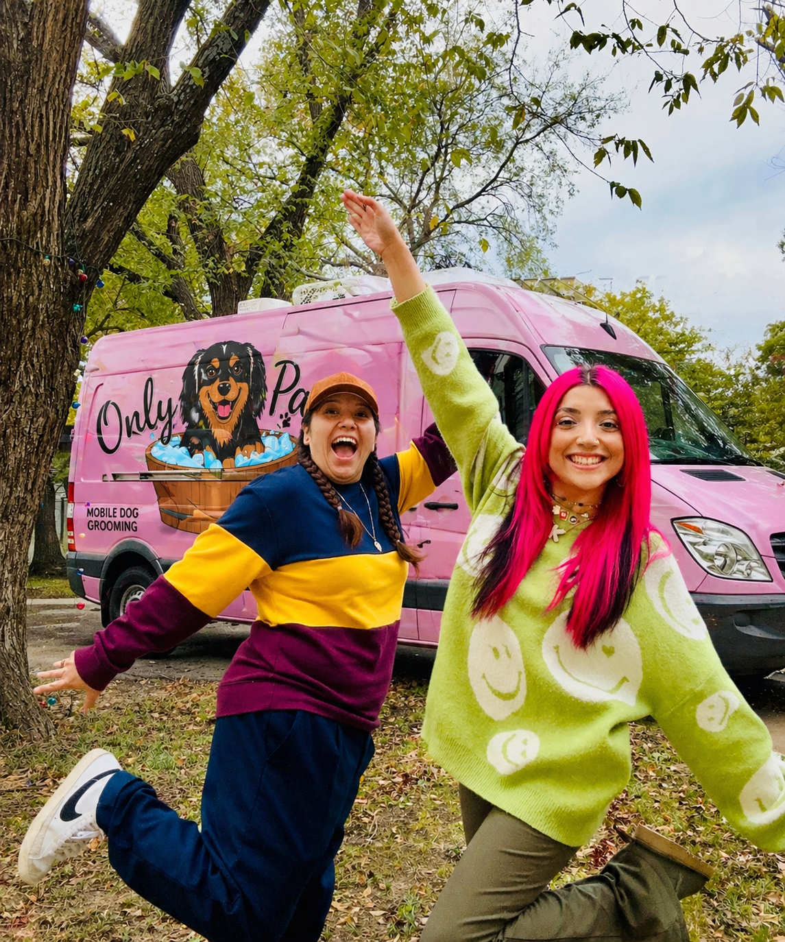 Two women smiling and playing joyfully outdoors in front of a pink mobile dog grooming van with a cartoon dog logo, surrounded by trees and autumn leaves.