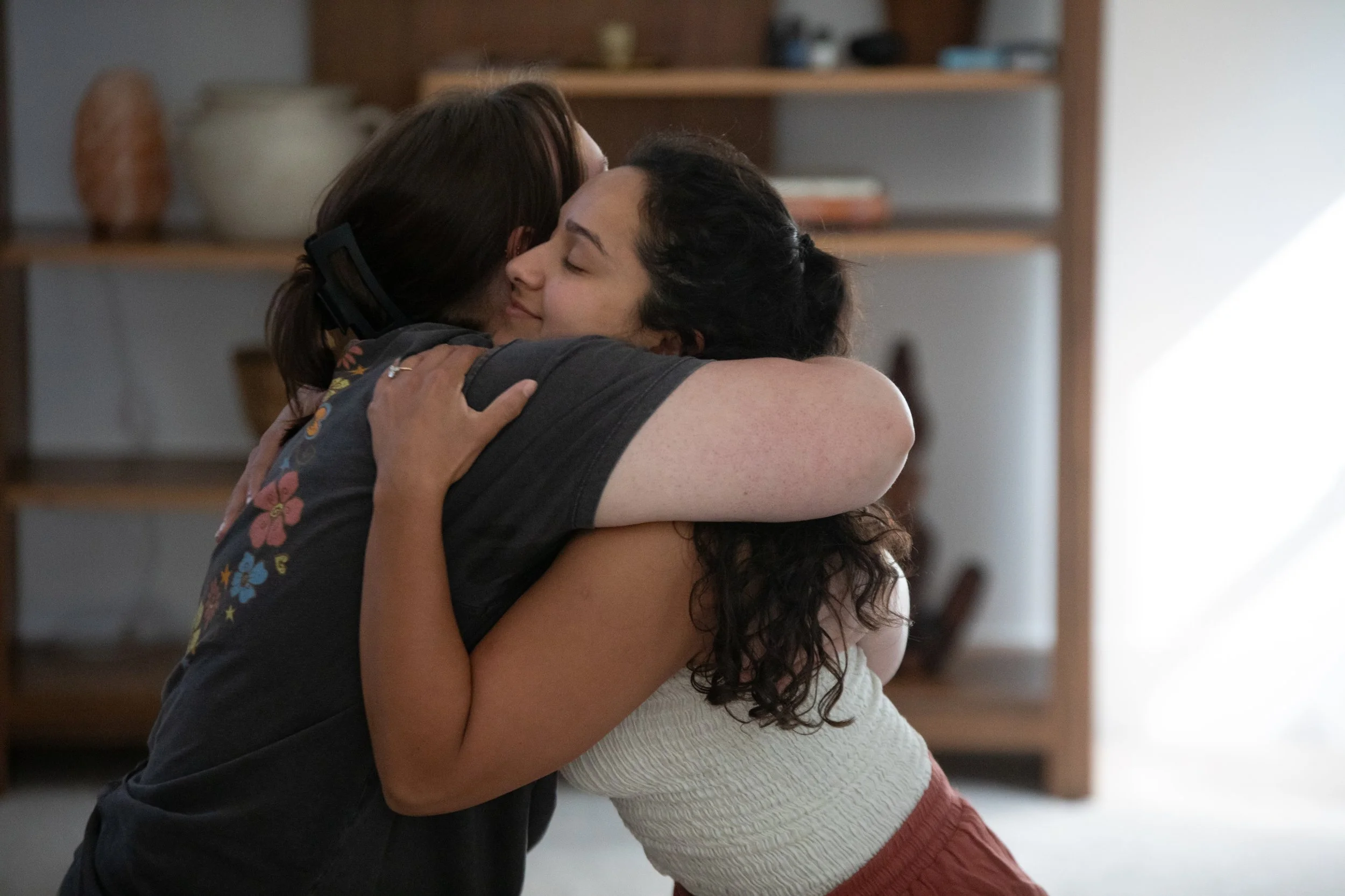Two women share an emotional hug in a cozy room, with blurred shelves and decorative items in the background.