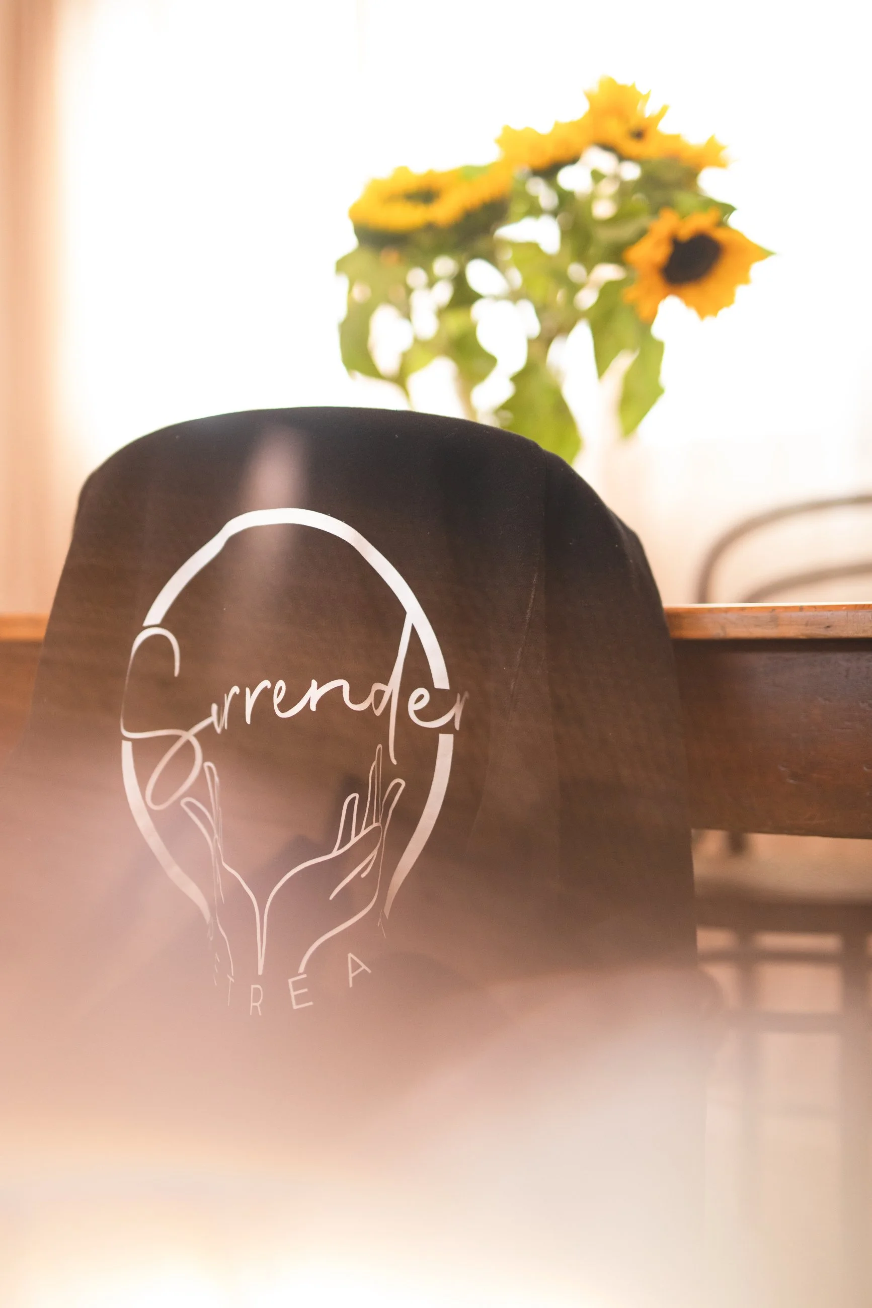 A black tote bag with the word "Surrender" and a graphic of two hands forming a heart shape, placed on a wooden table. In the background, a bouquet of sunflowers is visible.